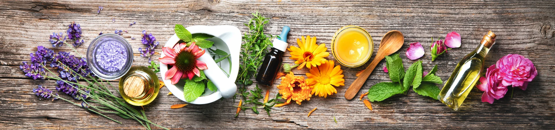 An arrangement of natural wellness items on a wooden surface includes a variety of colourful flowers, essential oil bottles, a bowl of lavender cream, a jar of honey, fresh herbs, and an aromatic oil dispenser.